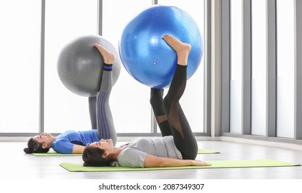 Two healthy Asian women practice yoga training and strength excercise by lying flat on their back on fitness mat, lifting legs up and nipping ball. Female adult work out for strong body at sport gym.