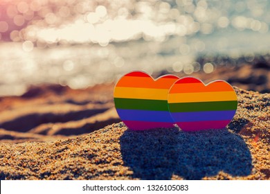 Two Hearts Of The Color Of The Gay Flag On Sand On The Coast Of The Sea. Romantic Trip Gay Honeymoon Same-sex Couple. LGBT Concept. Minority Sex. Heart Sign With Gay Pride LGBT Rainbow Flag Wristband.