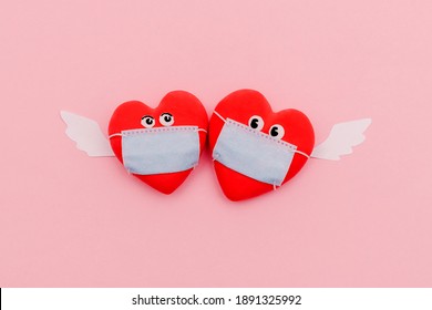 Two Hearts In Protective Masks With Eyes And Angel Wings On A Pink Background. Valentine's Day And Coronavirus Pandemic Concept.Flat Lay, Top View.