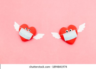 Two hearts in protective masks with eyes and angel wings on a pink background. Valentine's Day background. Coronavirus pandemic concept.Flat lay, top view.