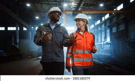 Two Heavy Industry Engineers In Hard Hats Walk In Steel Metal Manufacturing Factory And Have A Discussion. Black African American Industrial Specialist Talk To Female Technician In Manufacture.