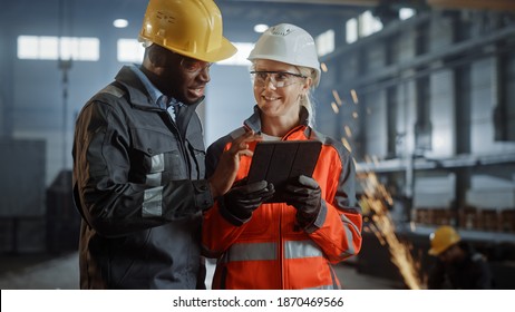 Two Heavy Industry Engineers Stand In Steel Metal Manufacturing Factory, Use Digital Tablet Computer And Have A Discussion. Black African American Industrial Specialist Talk To Female Technician.