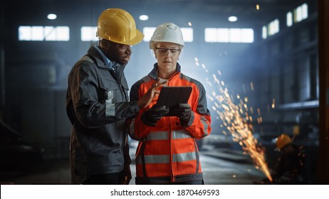 Two Heavy Industry Engineers Stand In Steel Metal Manufacturing Factory, Use Digital Tablet Computer And Have A Discussion. Black African American Industrial Specialist Talk To Female Technician.