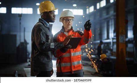 Two Heavy Industry Engineers Stand In Steel Metal Manufacturing Factory, Use Digital Tablet Computer And Have A Discussion. Black African American Industrial Specialist Talk To Female Technician.