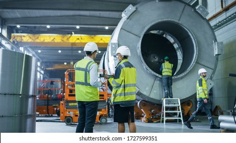 Two Heavy Industry Engineers Walk Through Pipe Manufacturing Factory, Use Digital Tablet Computer, Have Discussion. Facility For Construction Of Oil, Gas And Fuel Pipeline Transportation Products