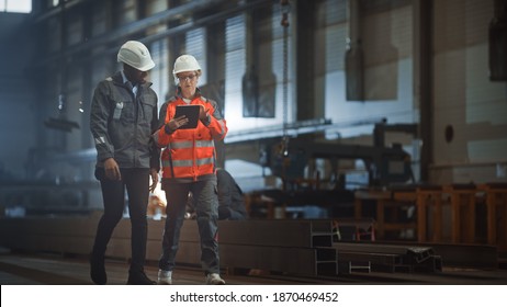 Two Heavy Industry Engineers Walk in Steel Factory, Use Tablet and Discuss Work. Industrial Worker Uses Angle Grinder in the Background. Black African American Specialist Talks to Female Technician. - Powered by Shutterstock