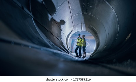 Two Heavy Industry Engineers Walking Inside Pipe, Use Laptop, Have Discussion, Checking Design. Construction Of The Oil, Natural Gas And Biofuels Transport Pipeline. Industrial Manufacturing Factory