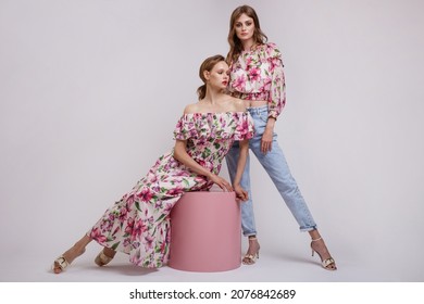 Two High Fashion Models In Long Dress With A Red Floral Pattern, Blouse, Blue Jeans. Beautiful Young Women. Studio Shot. White Background. Make Up, Hairstyle. Model Is Sitting On A Pink Cube Cylinder