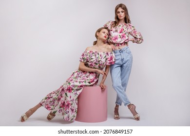 Two High Fashion Models In Long Dress With A Red Floral Pattern, Blouse, Blue Jeans. Beautiful Young Women. Studio Shot. White Background. Make Up, Hairstyle. Model Is Sitting On A Pink Cube Cylinder