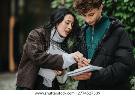 Two high school friends study from notebooks while talking and pointing at notes. A student pair reviews papers outdoors, collaborating on classwork and sharing study tips.
