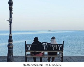 Two hijab women sitting on a bench by the waterfront, enjoying a peaceful moment while gazing at the calm sea under a clear sky, with a lamppost nearby - Powered by Shutterstock