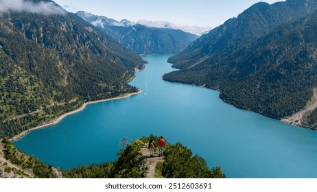 Two hikers enjoy the stunning views of Plansee Lake, framed by lush greenery and towering mountains under a clear sky. a couple of man and woman looking out over the lake - Powered by Shutterstock