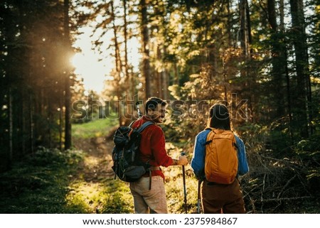 Two hikers, representing diversity, traverse a rugged terrain, their trusty backpacks packed with essentials as they navigate the natural wonders with their sturdy walking sticks