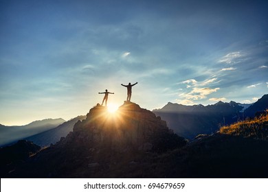 Два Туристов в силуэте стоит на скале в красивых горах с восходящими руками на фоне восхода неба