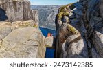 Two hikers sit on the edge of Kjeragbolten Pulpit Rock in Norway, enjoying the stunning panoramic view of the surrounding mountains and fjord. a couple of men and women at Kjeragbolten