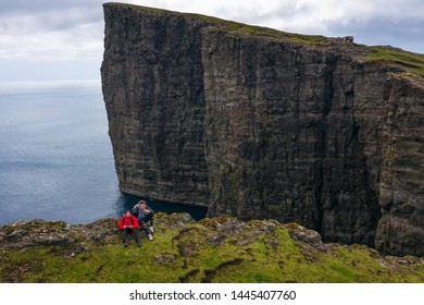 Two hikers sitting on the edge of a cliff with the famous Traelanipan cliff in the background. This cliff is located on the island of Vagar on Faroe Islands in Denmark. - Powered by Shutterstock