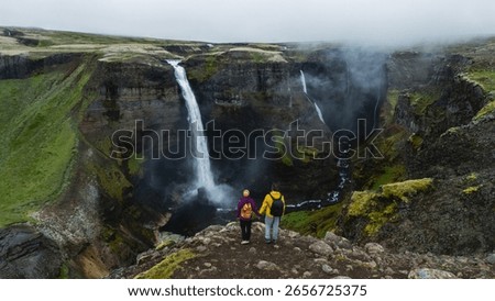 Two hikers stand at the edge of a dramatic cliff, gazing at the powerful Haifoss waterfall cascading into a lush valley below in Iceland