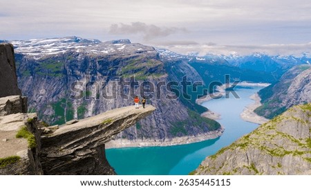 Two hikers stand on a dramatic cliff edge in Trolltunga, Norway, taking in breathtaking views of the turquoise fjord below. Majestic mountains surround the captivating landscape under a cloudy sky.