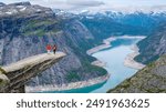 Two hikers stand on a rocky precipice overlooking a picturesque Norwegian fjord, with snow-capped mountains in the distance. Trolltunga, Norway,