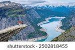 Two hikers stand on a rocky precipice overlooking a picturesque Norwegian fjord, with snow-capped mountains in the distance. Trolltunga, Norway,