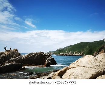 Two hikers stand on rocky cliffs overlooking the ocean with a backdrop of green hills and a clear blue sky. - Powered by Shutterstock