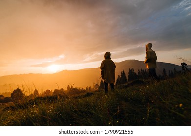 Two hikers stand in the rain on a meadow in the mountains and watch the incredible orange sunset. Two hikers in raincoats and sunset in the rain on a background of beautiful landscape. - Powered by Shutterstock