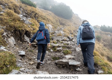 Two hikers wearing backpacks trekking uphill on rocky mountain trail surrounded by mist and fog. Concept of outdoor adventure, hiking in nature, enjoying active lifestyles in all-weather conditions. - Powered by Shutterstock