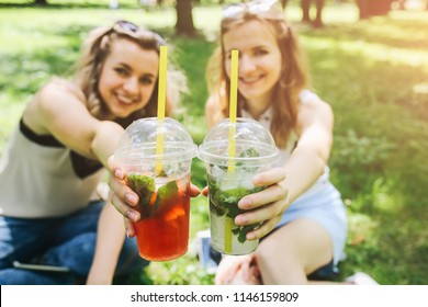 Two Hipster Girls Are Laughing And Drinking Summer Cocktails Outdoors In The Green Grass. Cold Non-alcoholic Drinks With Ice To Go. Mojito And Strawberry Lemonade. Happy Lifestyle For Vacations.