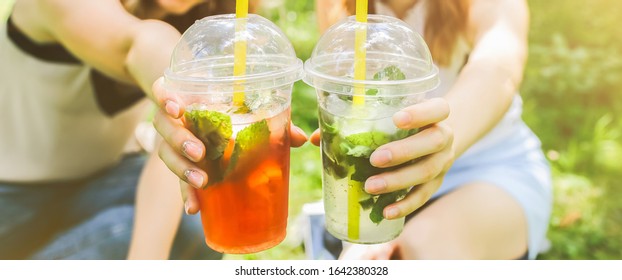 Two Hipster Girls Are Laughing And Drinking Summer Cocktails Outdoors In The Green Grass. Cold Non-alcoholic Drinks With Ice To Go. Mojito And Strawberry Lemonade. Happy Lifestyle For Vacations.