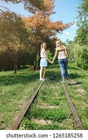 Two Hipster Girls Are Walking On The Railway In The Green Park. Same Sex Couple Is Holding Hands Together. Travel Style Life Concept. Support And Friendship Relationships. Spring Time.