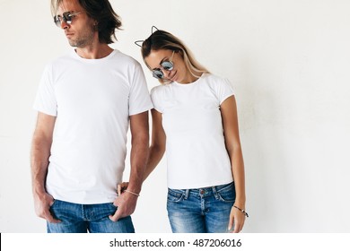 Two Hipster Models Man And Woman Wearing Blanc T-shirt, Jeans And Sunglasses Posing Against White Wall, Toned Photo, Front Tshirt Mockup For Couple