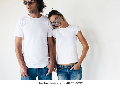 Two hipster models man and woman wearing blanc t-shirt, jeans and sunglasses posing against white wall, toned photo, front tshirt mockup for couple