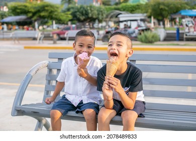 Two Hispanic Cheerful Kids Sitting On A Bench In A Park And Eating Ice Cream