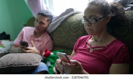 Two Hispanic Family Members Sitting On Couch Looking At Cellphones. A Senior Woman Looking At Phone Screen With Grandson