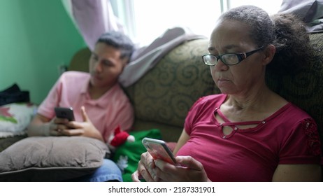 Two Hispanic Family Members Sitting On Couch Looking At Cellphones. A Senior Woman Looking At Phone Screen With Grandson