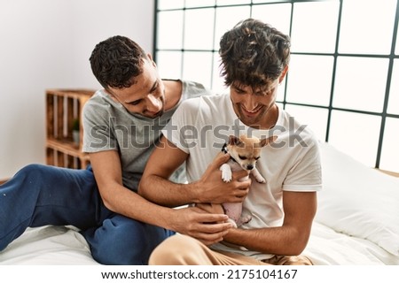Two hispanic men couple hugging each other sitting on bed with chihuahua at bedroom