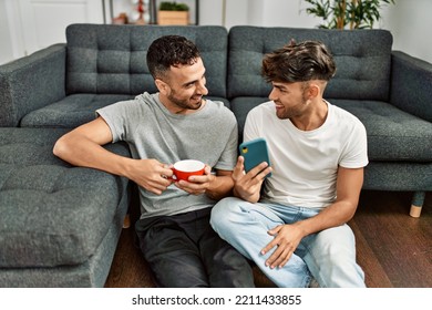 Two hispanic men couple using smartphone and drinking coffee at home