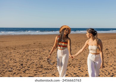 Two hispanic multicultural lesbian girls walk hand in hand, expressing their love as they stroll along the beach on summer day, showcasing the beauty of LGBTQ+ relationships amidst the coastal scenery - Powered by Shutterstock