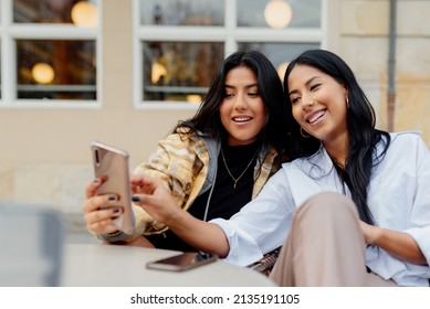 Two Hispanic Sisters Taking A Picture With Their Smart Phone In The Street. Young Latin Girls Using A Mobile Phone And Having Fun Happily.