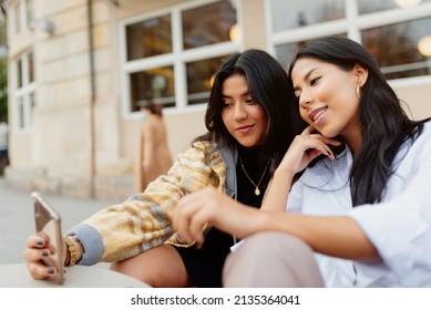 Two Hispanic Sisters Taking A Picture With Their Smart Phone In The Street. Young Latin Girls Using A Mobile Phone And Having Fun Happily.