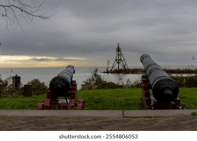 Two historic cannons facing the sea with a cloudy sky and a beacon in the distance, capturing a blend of maritime heritage and serene coastal scenery. - Powered by Shutterstock