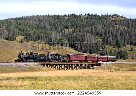 Two Historic Steam Locomotives Pulling an Antique Wooden Passenger Train Over a Railroad Bridge in a Scenic Meadow with a Mountain Forest in the Background