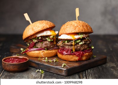 Two homemade beef burgers with mushrooms, micro greens, red onion, fried eggs and beet sauce on wooden cutting board. Side view, close up - Shutterstock ID 1009968298