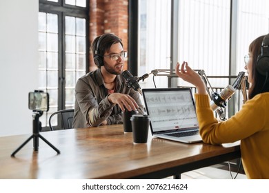 Two hosts man and woman podcasting together, talking while shooting video podcast on smartphone in broadcasting studio. Woman radio hosts gesturing to microphone while interviewing man guest - Powered by Shutterstock