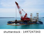 two huge excavators together synchronously on floating platforms in the blue water of the Pacific Ocean extract white sand from the bottom and load onto a barge amid clouds and tropical islands