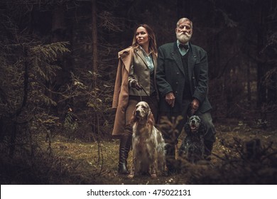 Two hunters with dogs and shotguns in a traditional shooting clothing on a dark forest background.