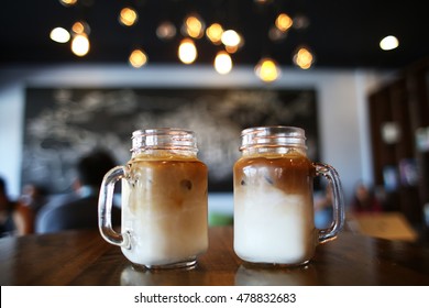 Two Iced Latte Coffees In Glass Mug Jar With Twinkle Lights In Background On Wooden Table At Cafe