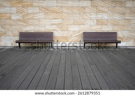 Two identical empty park benches positioned on a wooden surface in front of a sandstone wall.