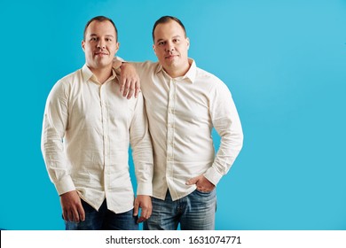 Two Identical Male Twins Wearing White Shirts And Jeans Standing Against Blue Background Looking At Camera