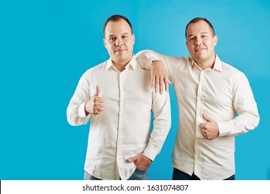 Two Identical Male Twins Wearing White Shirts Standing Against Blue Background Looking At Camera Showing Thumbs Up